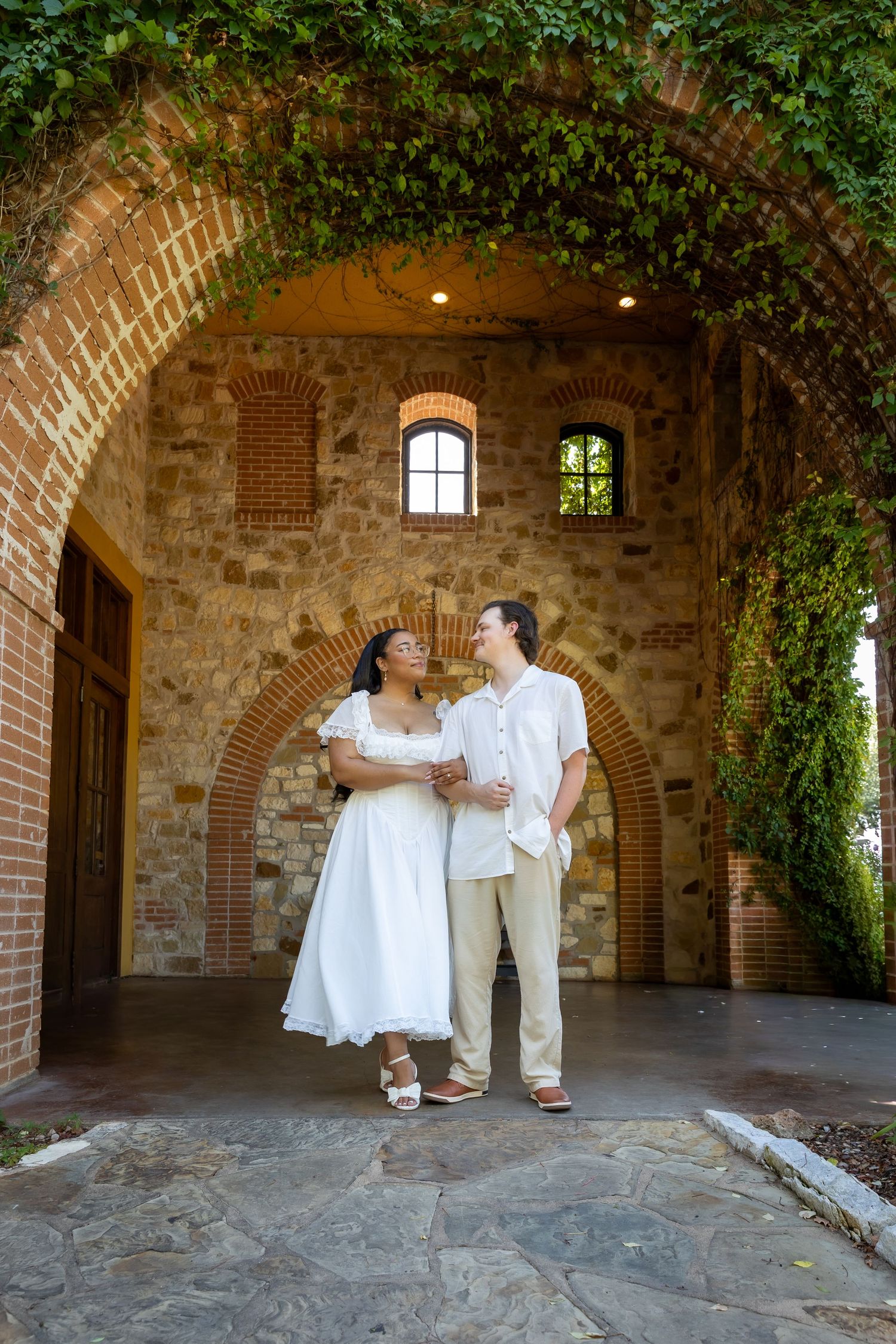 A bride and groom share a romantic kiss outdoors, with the bride in a white lace gown and groom in a tan suit.
