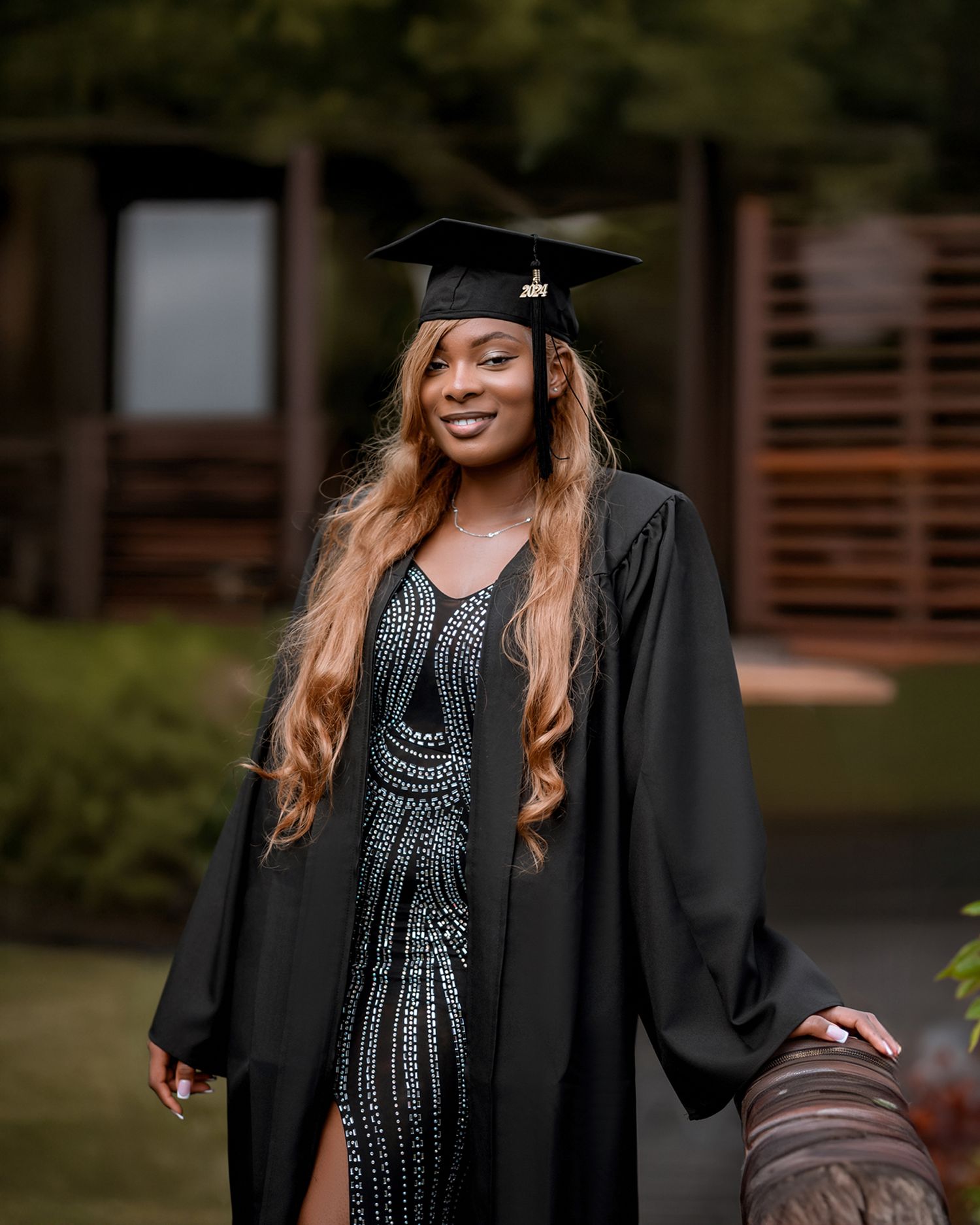 A smiling graduate in black academic gown sits on a wooden bench holding red roses outdoors on a sunny day.
