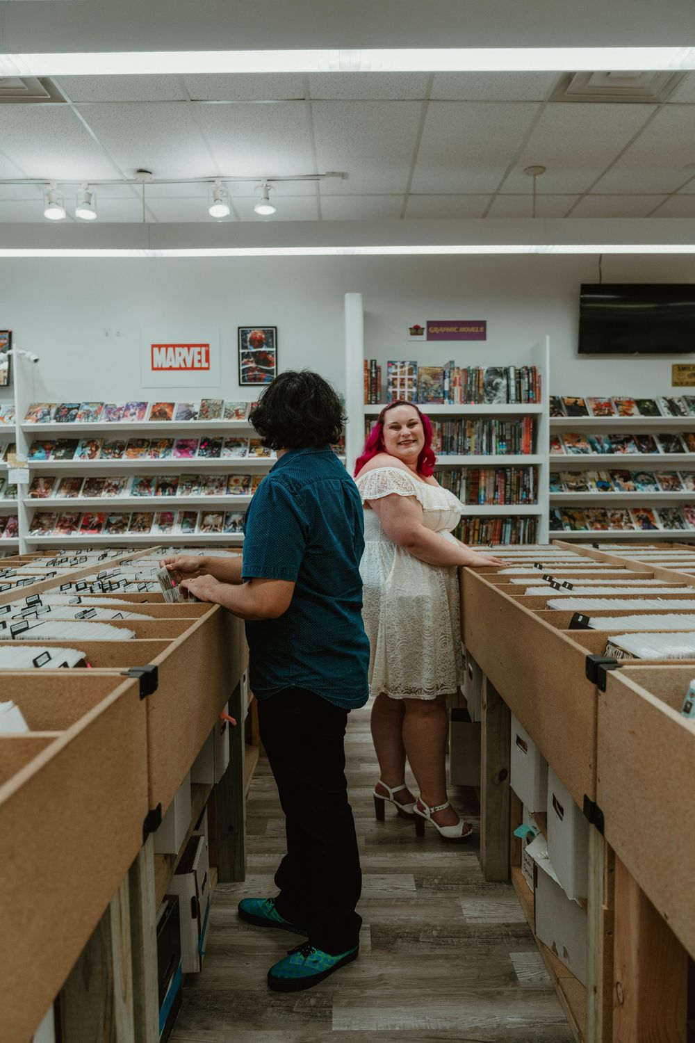 Couple browsing comic book store aisles, woman with pink hair in white dress smiling at camera.