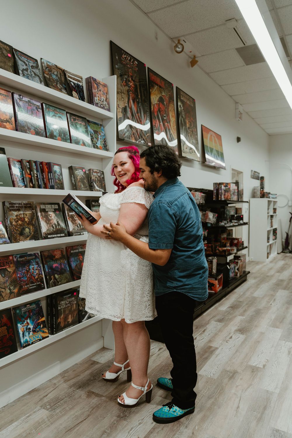 Couple embracing in a comic book store, woman in white lace dress with pink hair holding a book.