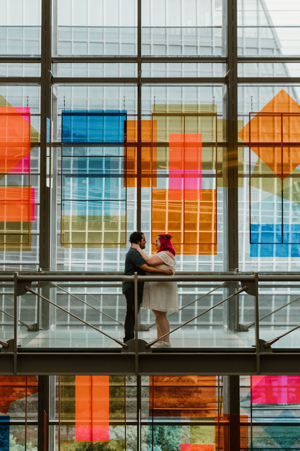 Couple embraces on a balcony surrounded by colorful geometric glass panels in a modern building.