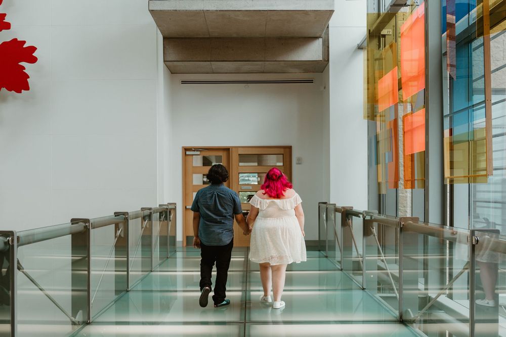 Two people walking down a glass-floored corridor toward wooden doors in a modern building with colorful windows.