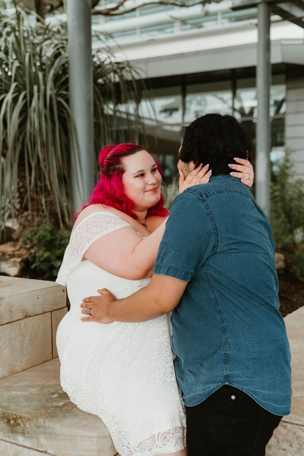 A woman with pink hair in a white lace dress embraces her partner outdoors in a tender romantic moment.