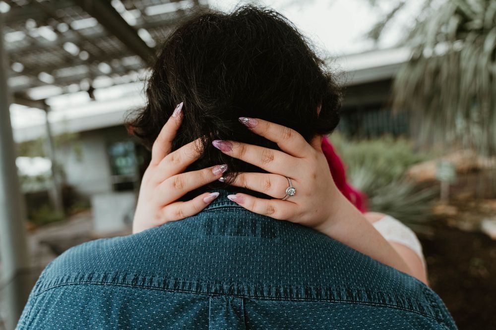 A woman with purple nails and a diamond ring embraces a man in a denim shirt from behind outdoors.