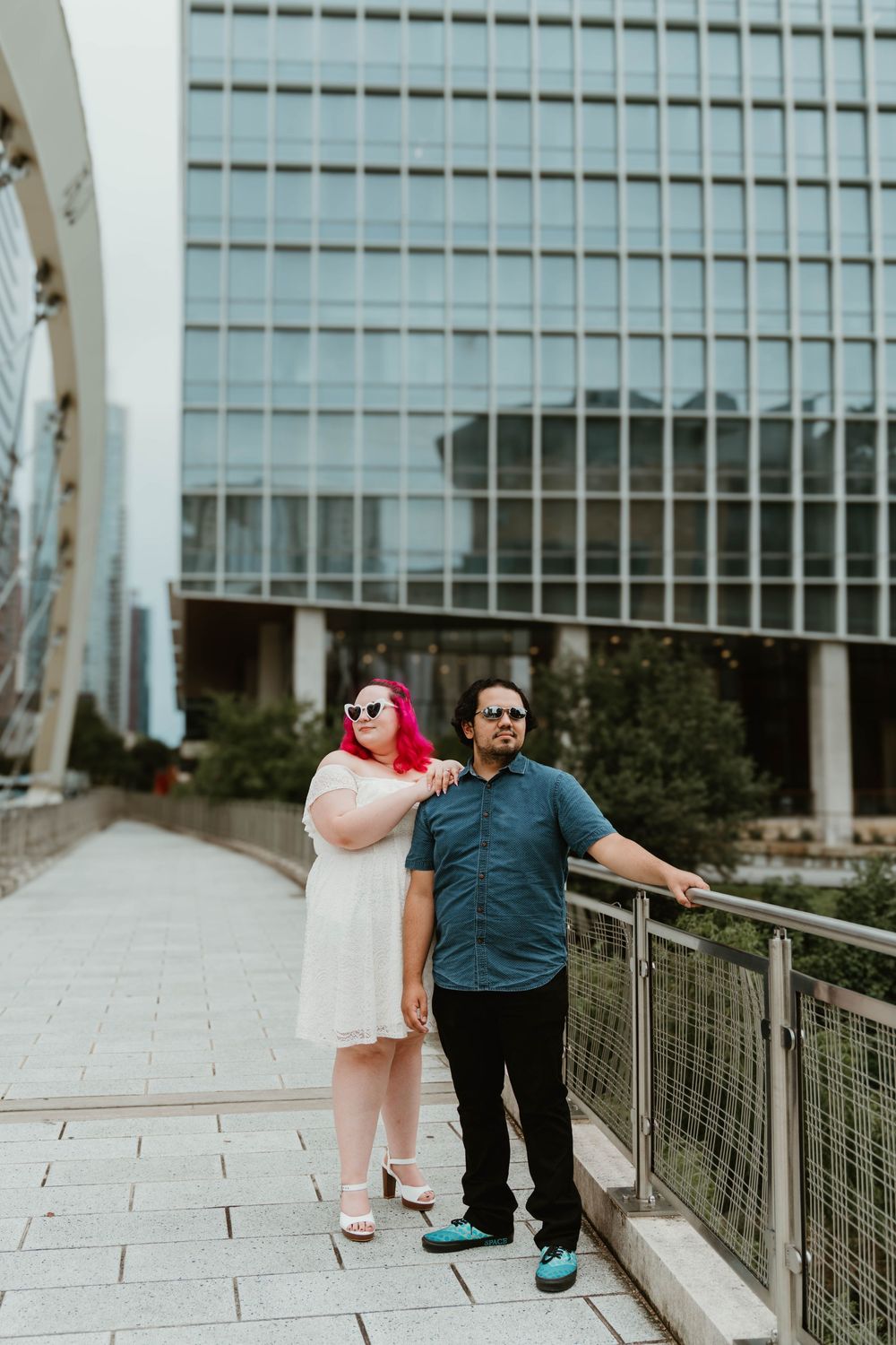 Couple posing on urban walkway, woman with pink hair in white dress, man in teal shirt, modern glass building backdrop.