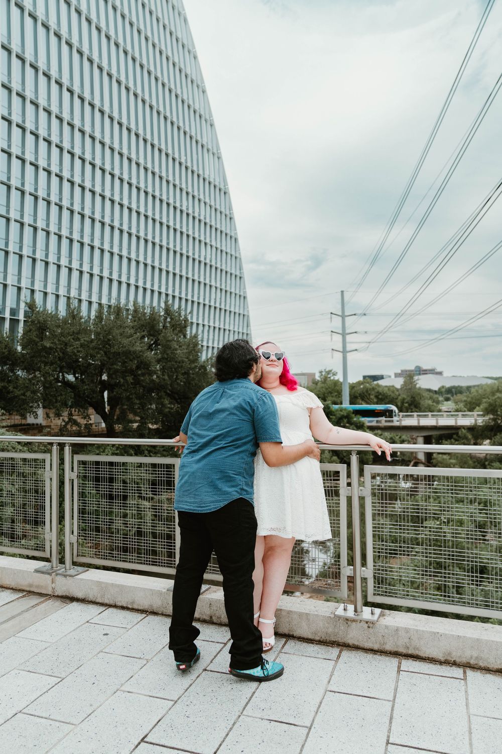 Man kissing woman with pink hair in white dress on city bridge, glass skyscraper in background.