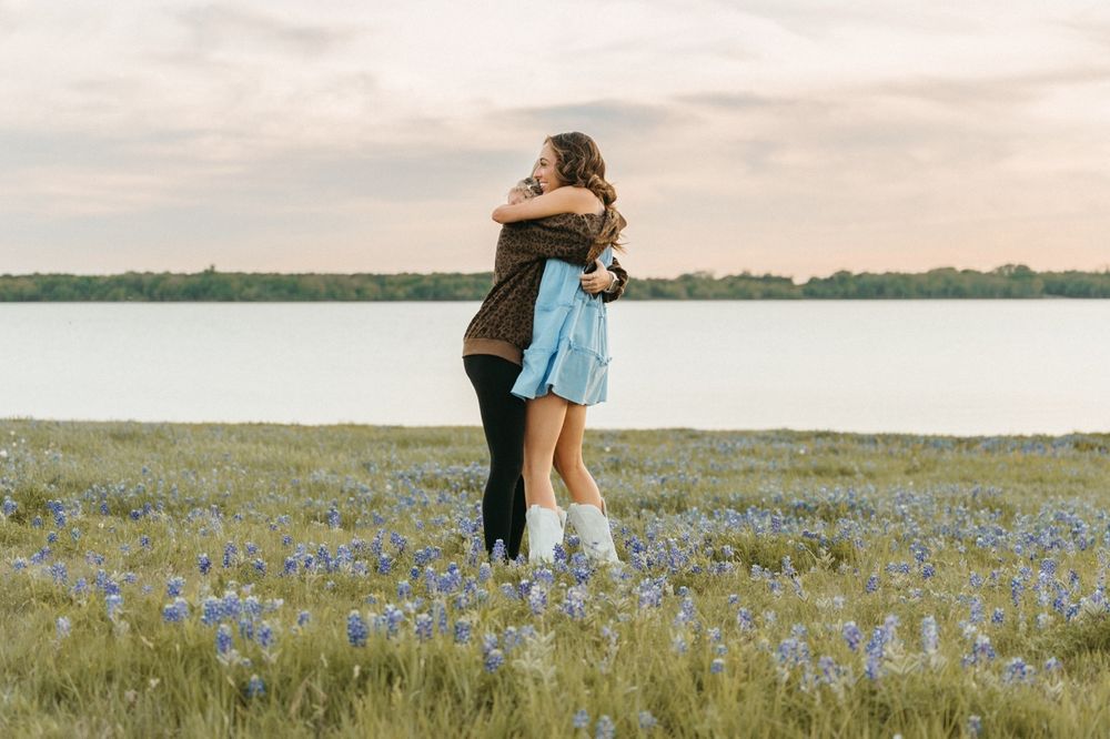 Couple embracing in a bluebonnet field by a lake at sunset.
