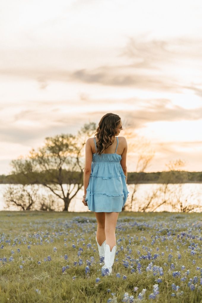 Young woman in blue ruffled dress and white cowboy boots standing in bluebonnet field at sunset.