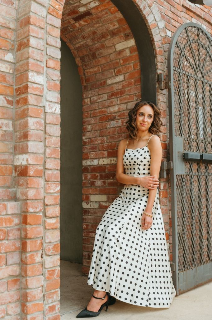 Woman in white polka dot midi dress posing in brick archway doorway with black heels.