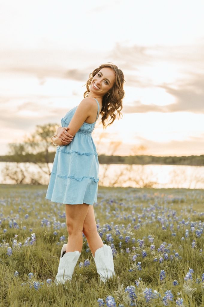Young woman in blue dress and white cowboy boots smiling in a field of bluebonnets at sunset.