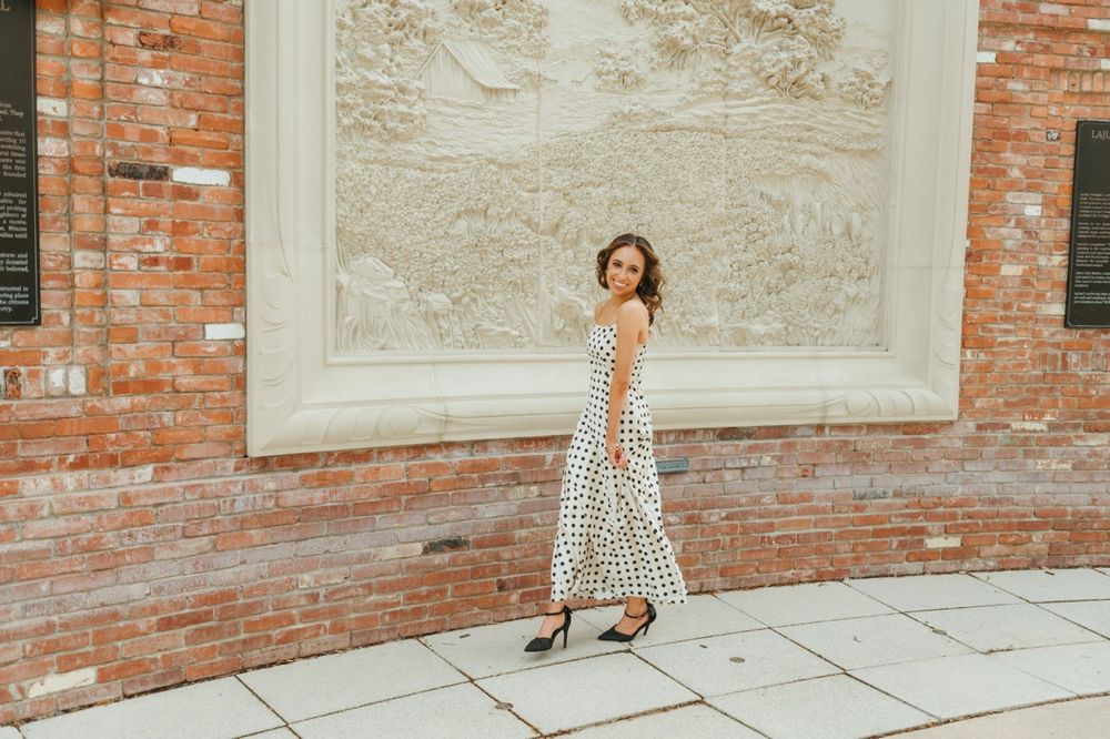 Woman walking in white polka dot dress past brick wall with decorative stone relief panel.