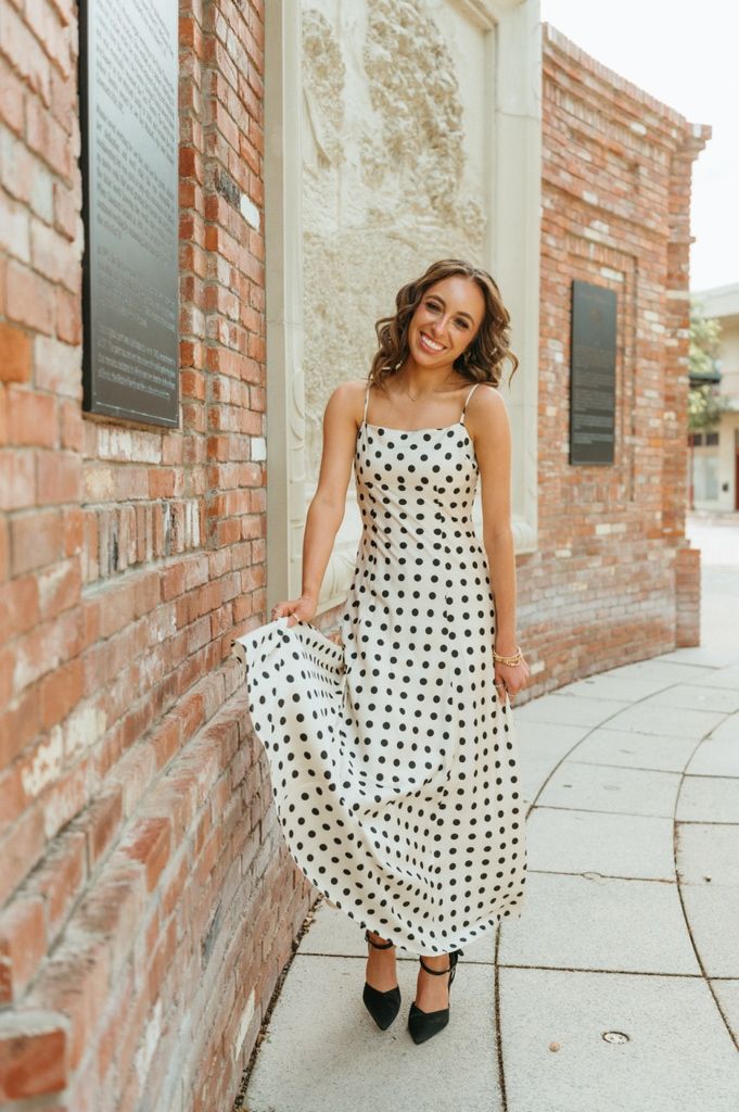 Woman in white polka dot maxi dress with spaghetti straps and black heels posing on a brick sidewalk.