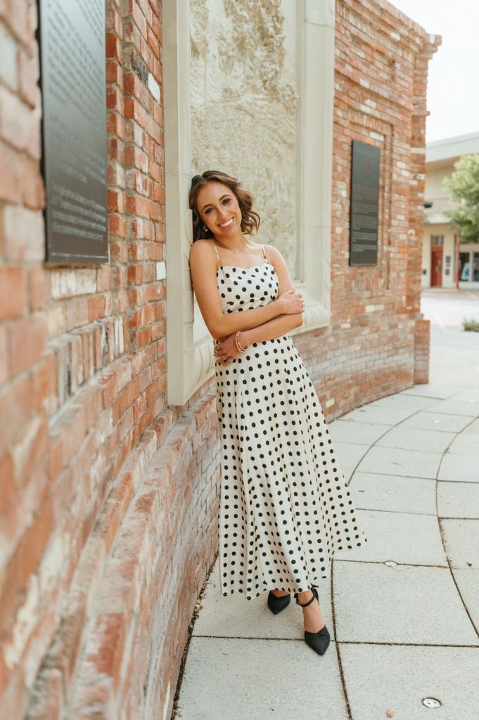 Woman in white polka dot maxi dress leaning against a brick wall with arms crossed wearing black heels.