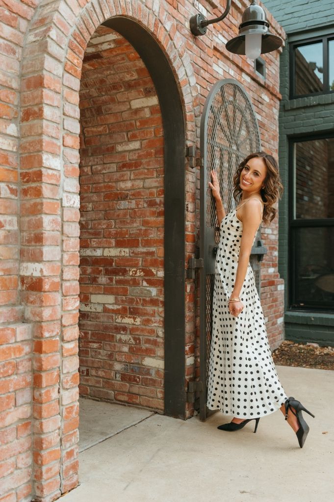 Woman in white polka dot midi dress and black heels posing by rustic brick archway door.