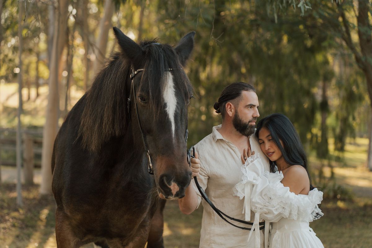 A couple in white attire poses with a dark horse outdoors among trees in a romantic photo shoot in sydney