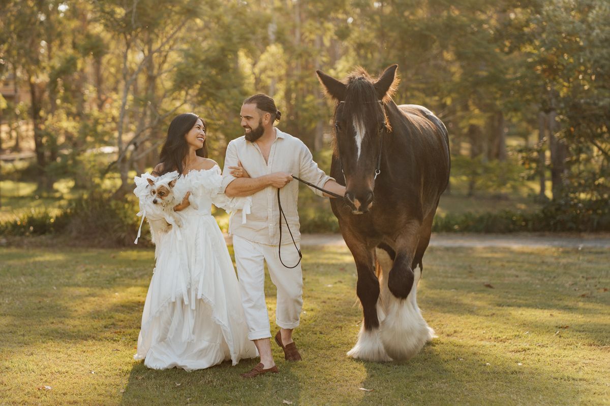 Couple in white attire walking with a large brown horse in a lush green outdoor setting during a photoshoot with a horse in sydney