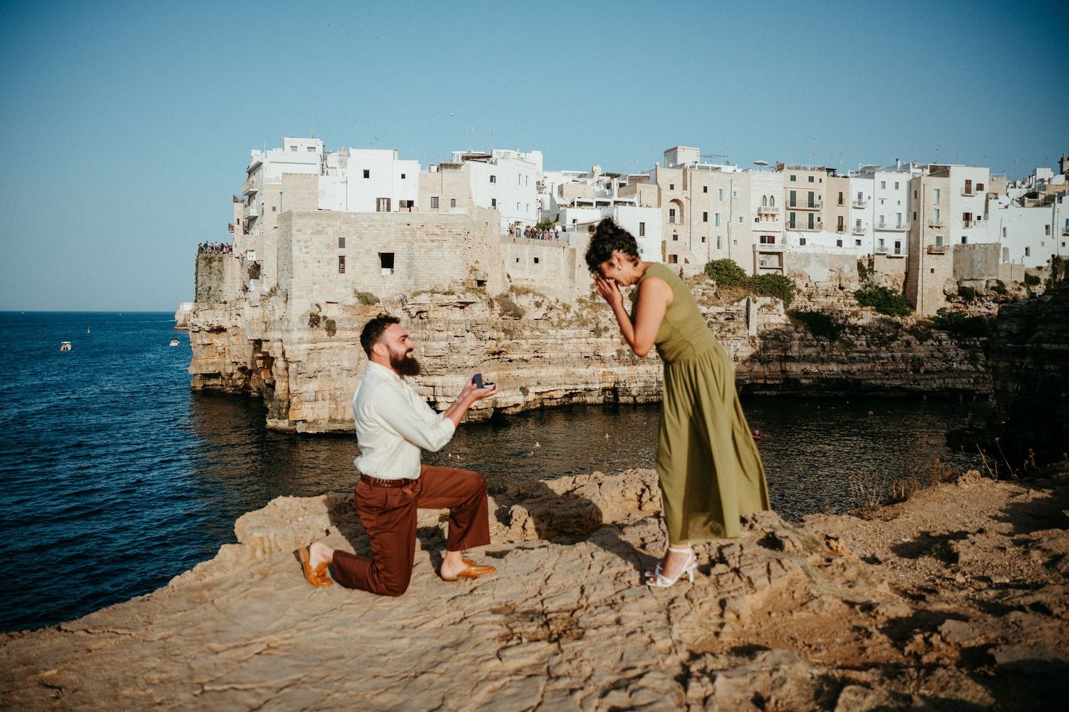 Man proposing to woman on rocky cliffside overlooking white coastal Italian village of Polignano a Mare.