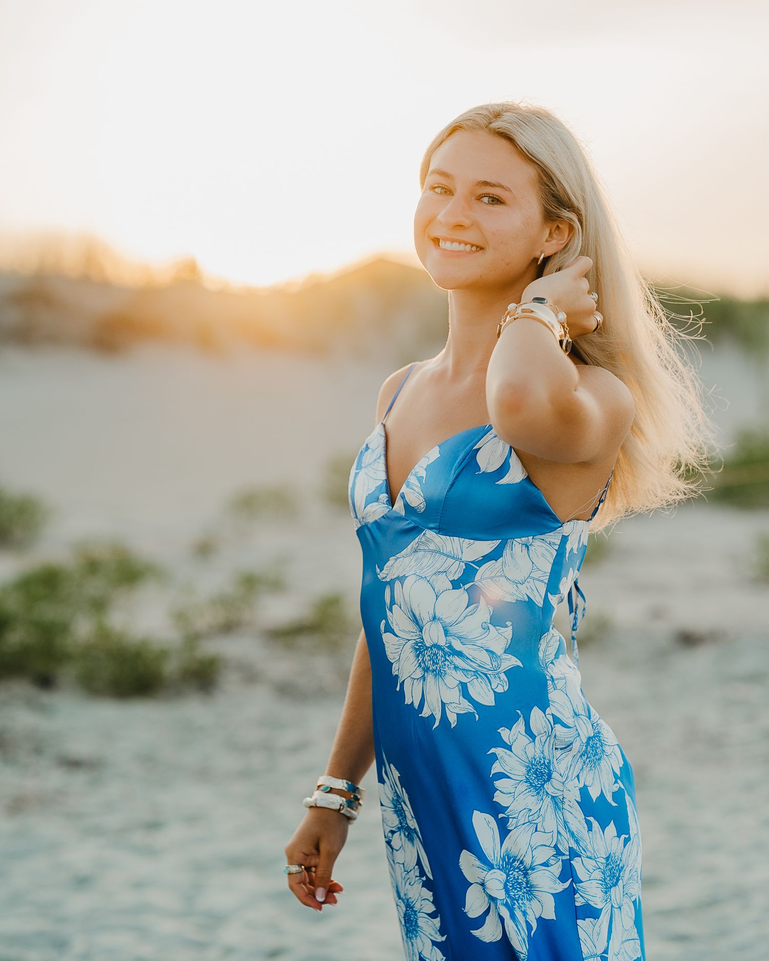 Young woman in a blush sundress leaning against a wooden post on a sandy beach, smiling warmly.