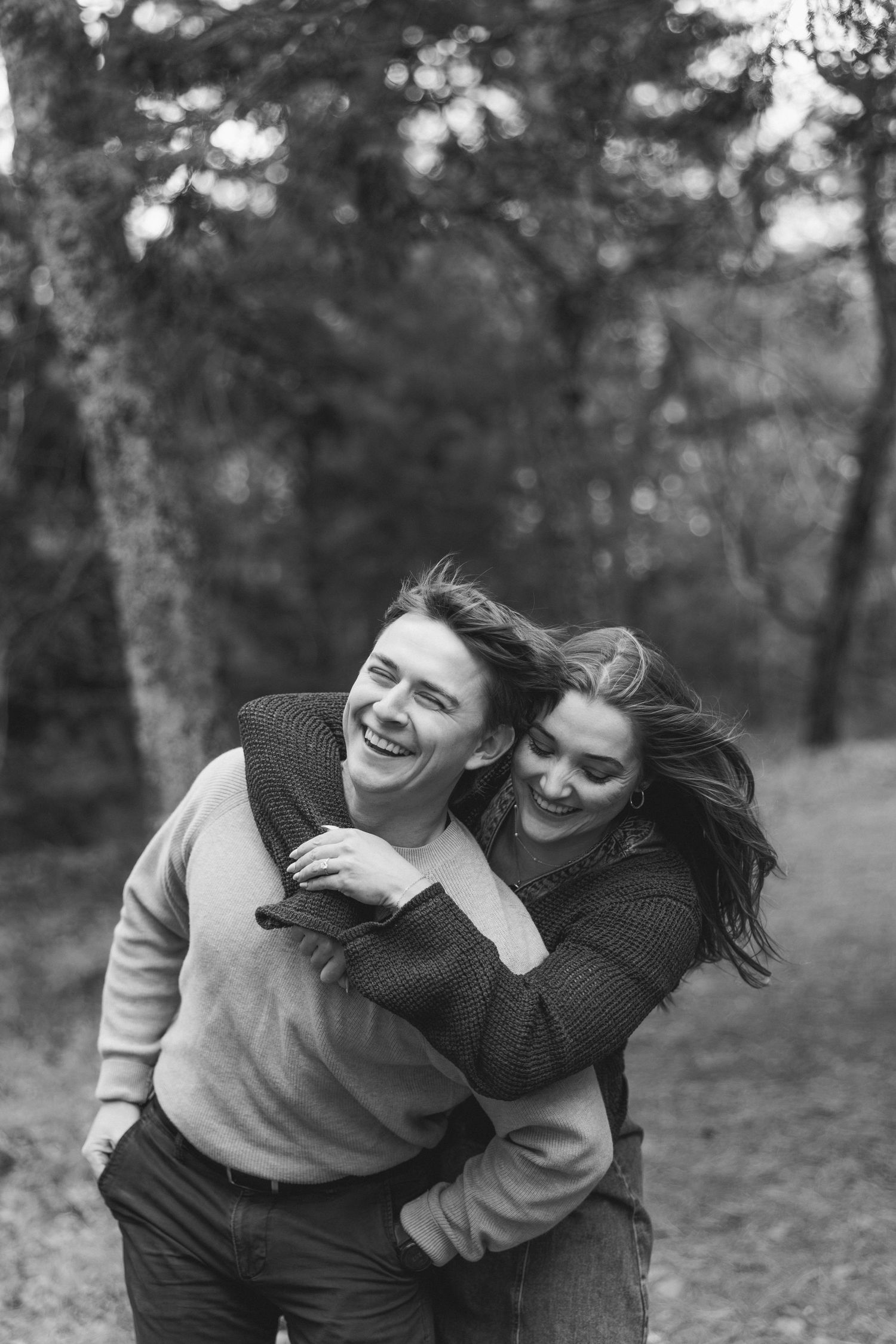 Black and white photo of a laughing couple embracing outdoors in a forest, woman hugging man from behind.
