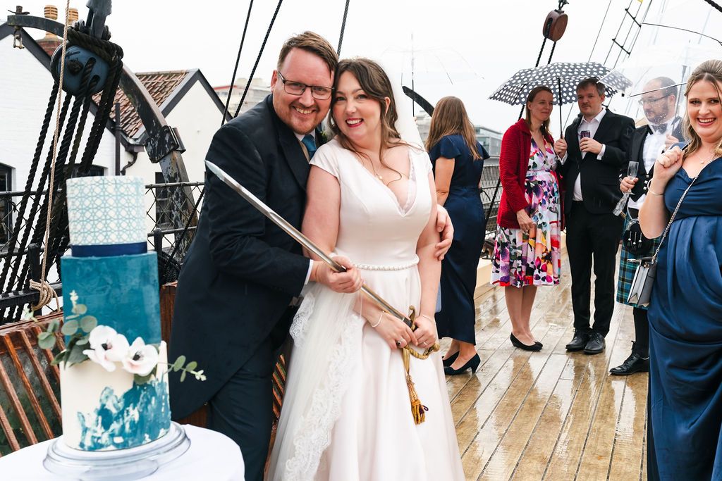 Bride and groom cutting wedding cake with a sword on a ship deck, guests with umbrellas in background.