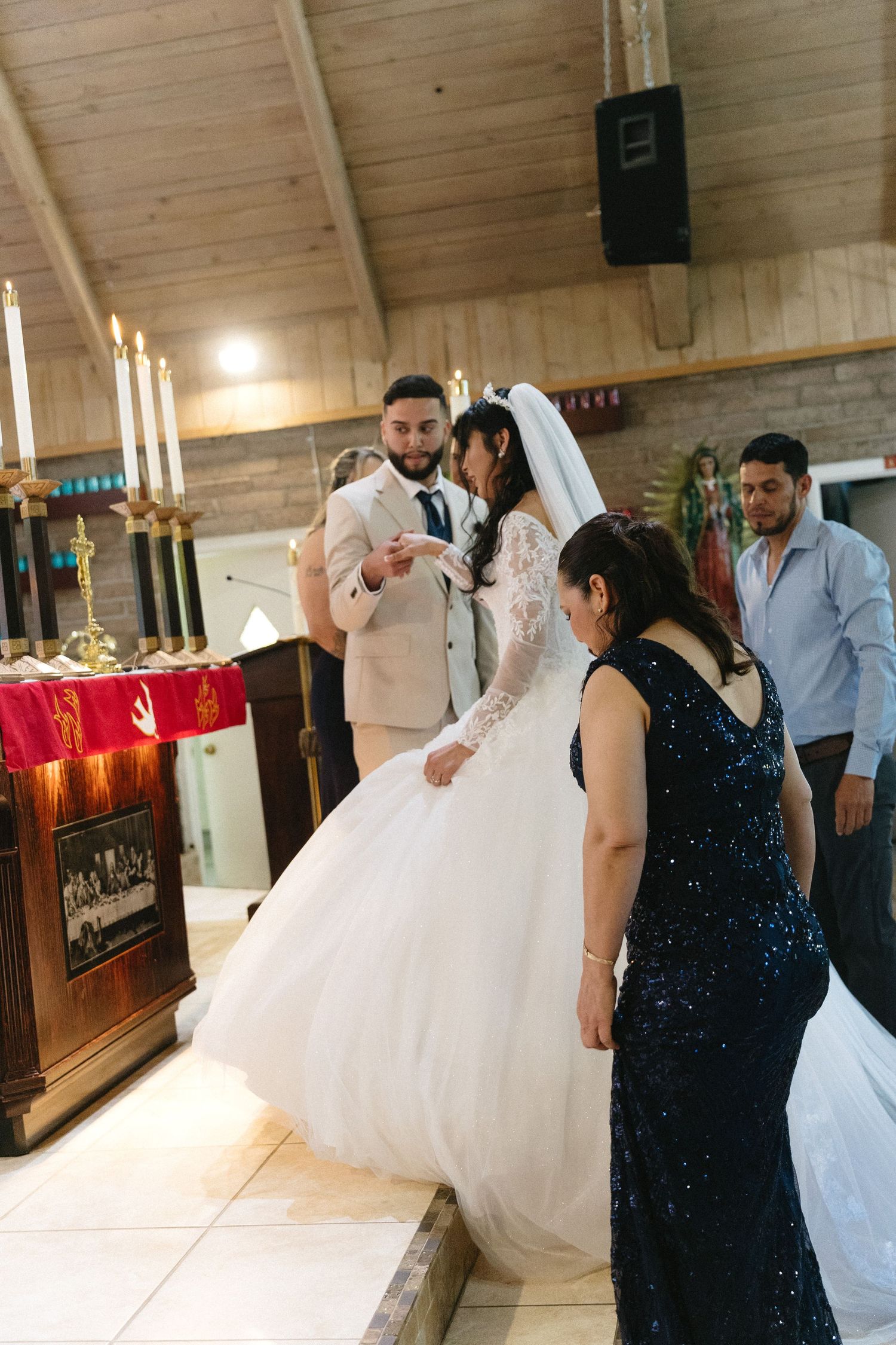 A bride in a white veil stands at the altar with a groom in a beige suit, as a woman in a navy sequin gown looks on.