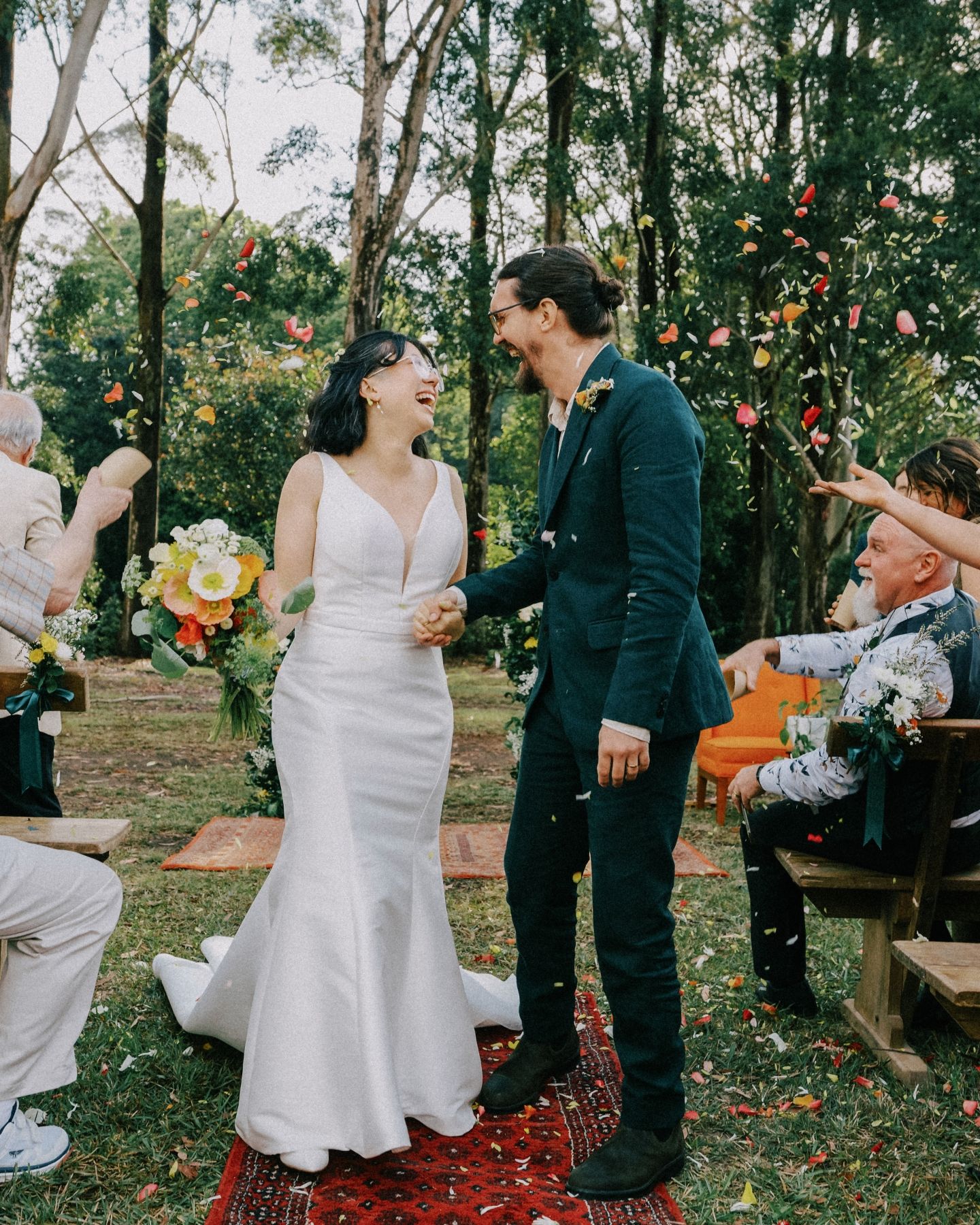 A joyful bride and groom walk down the aisle outdoors as guests toss colorful flower petals at their woodland wedding.
