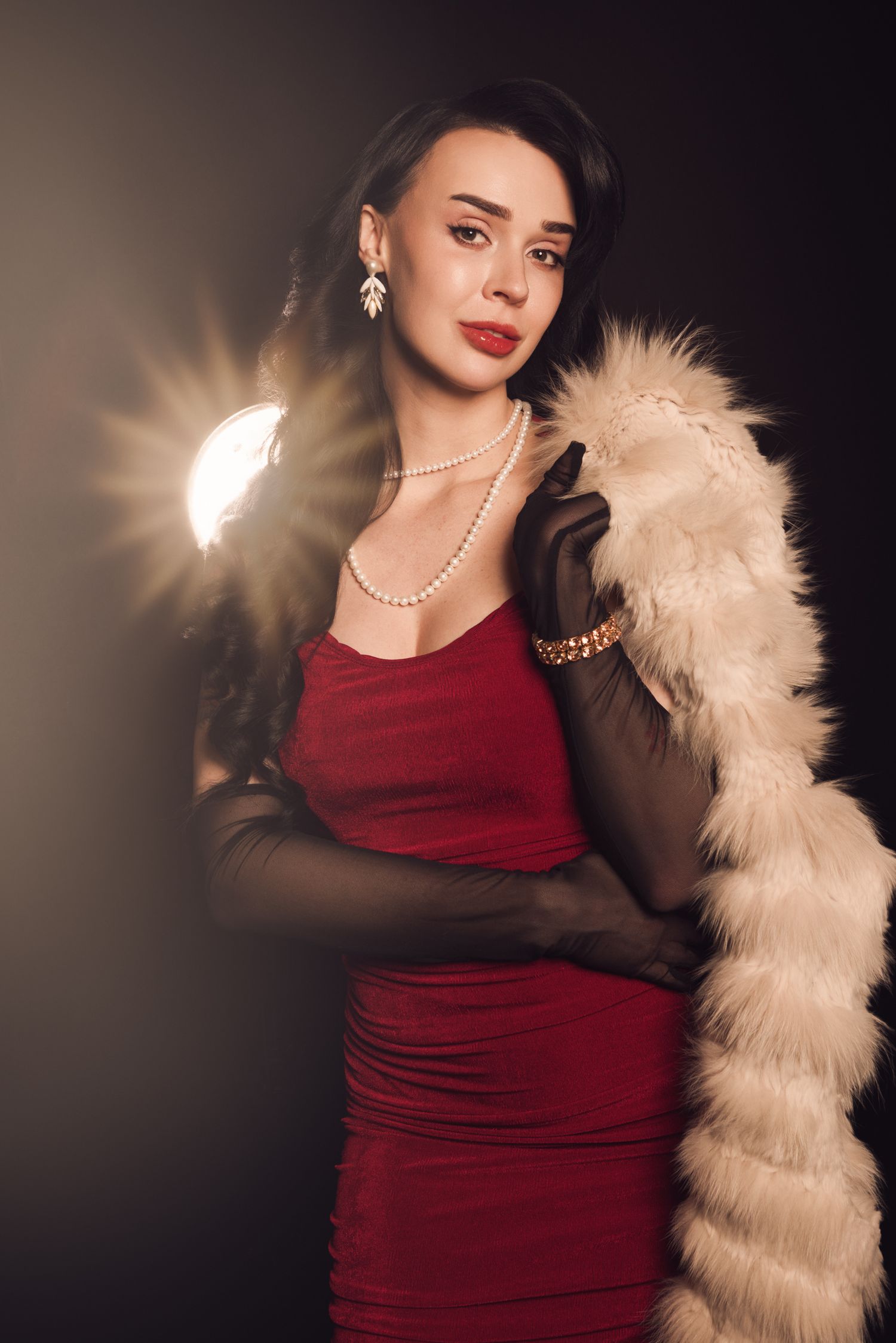 A smiling Black woman in a mauve halter dress drapes a white fur coat over her shoulder against a dark studio backdrop.