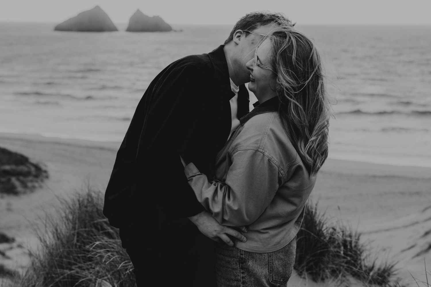 Couple sharing a romantic kiss in the dunes at Holywell Bay in black and white photography with ocean backdrop.