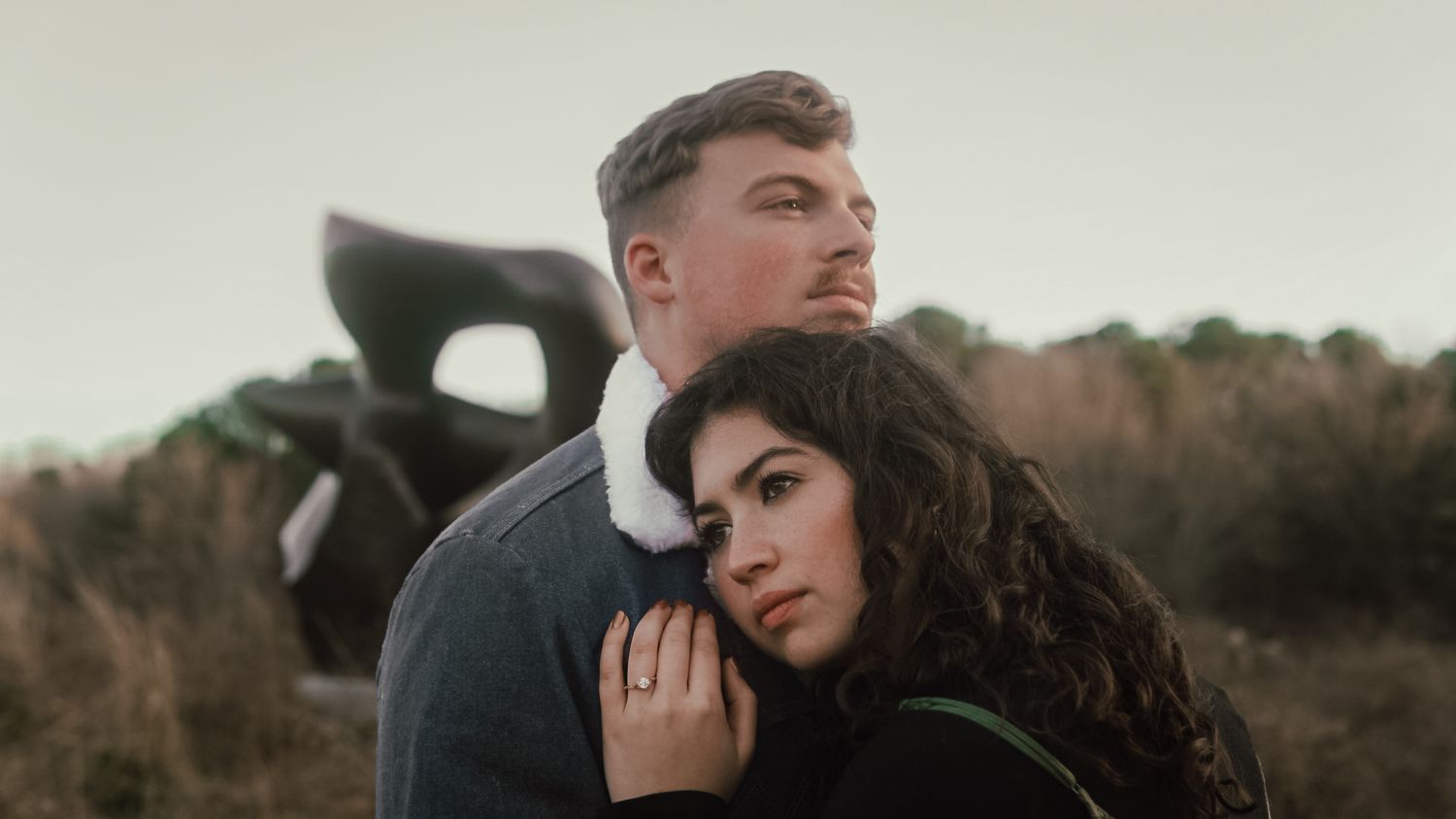 Young couple posing outdoors in a field, woman leaning back against man with sculpture behind.