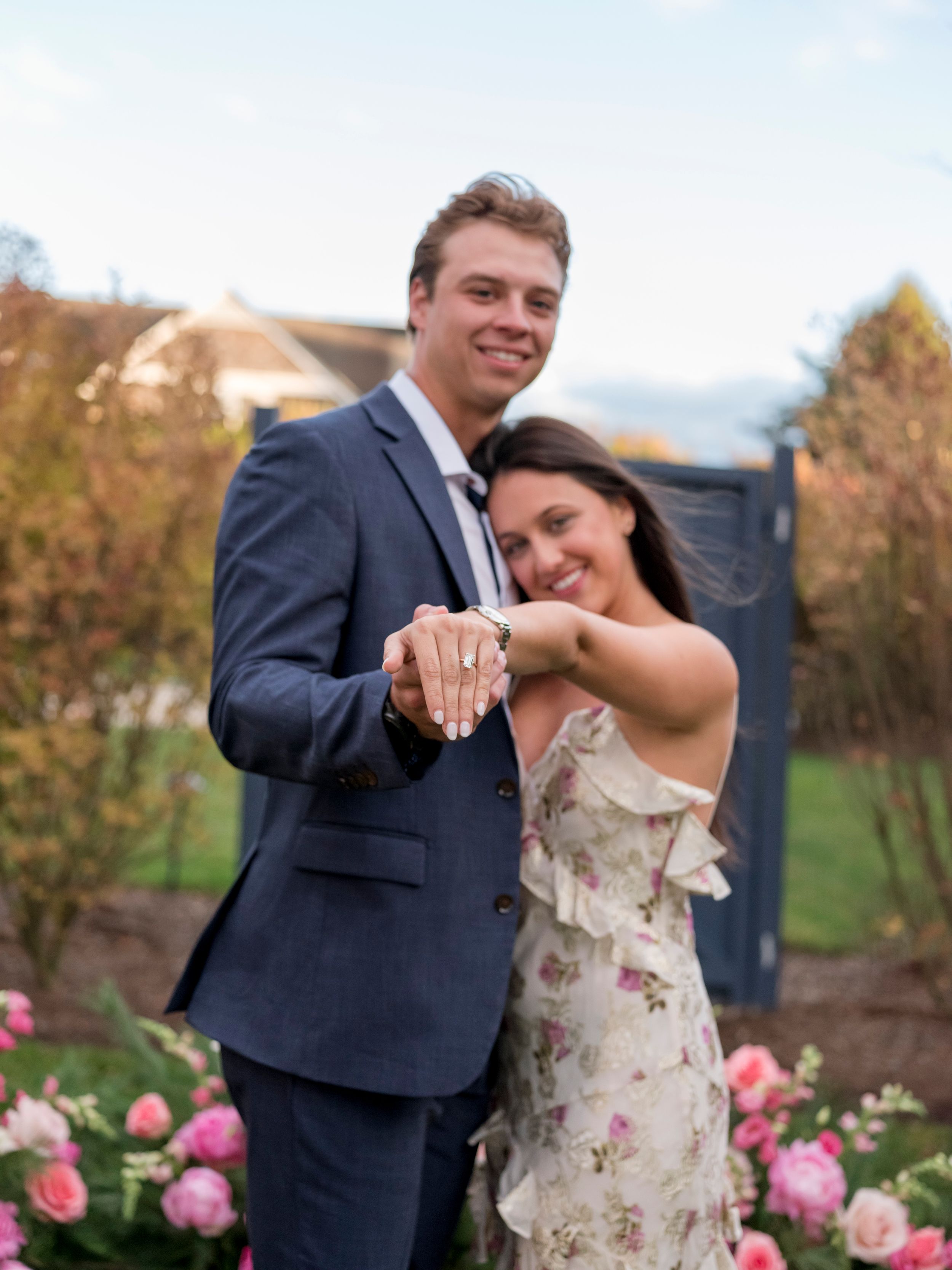Happy couple smiling and showing off engagement ring outdoors among pink flowers, man in navy suit, woman in floral dress.