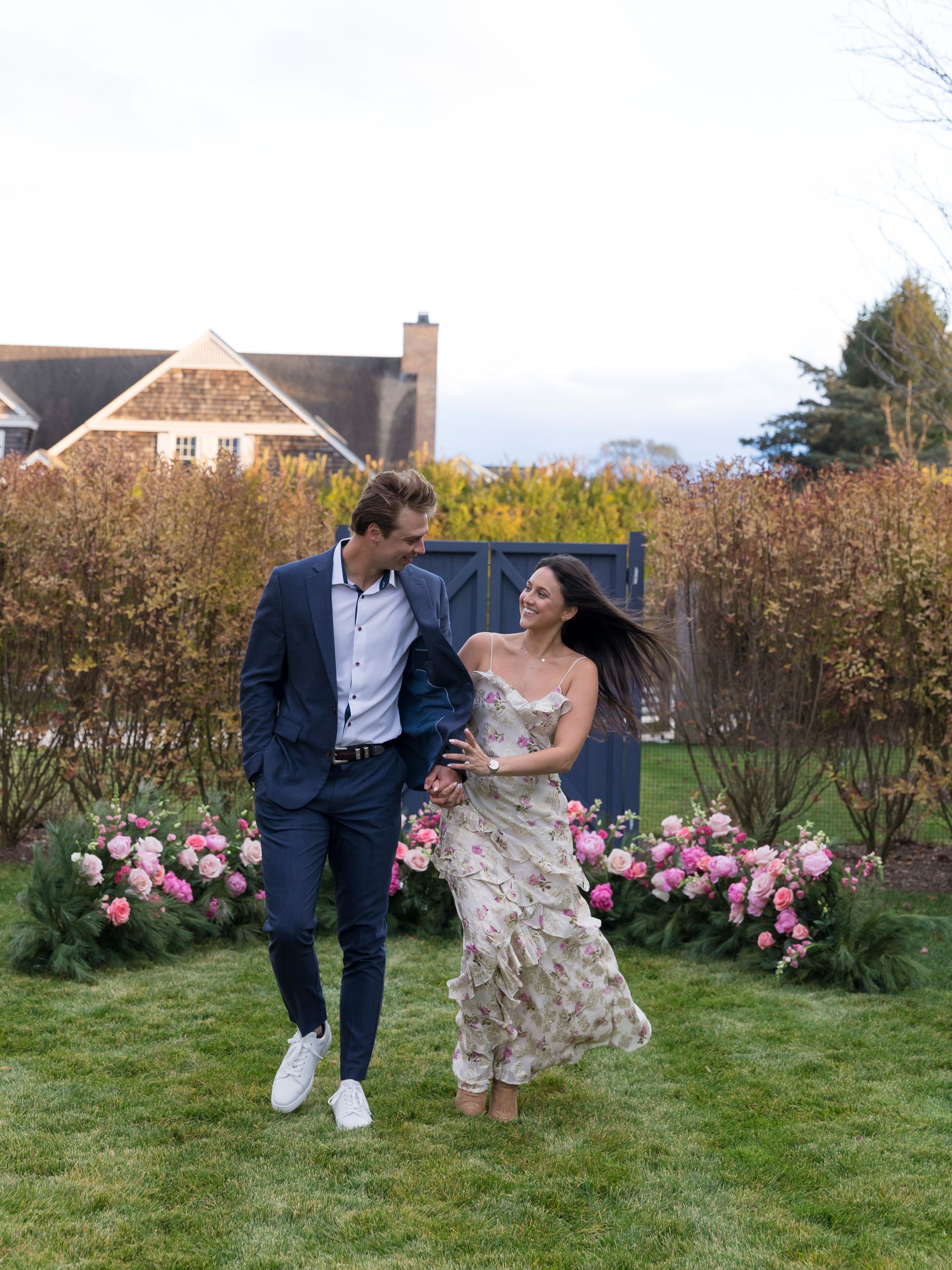 Couple laughing and walking together in a garden with pink roses, man in navy suit, woman in floral dress.