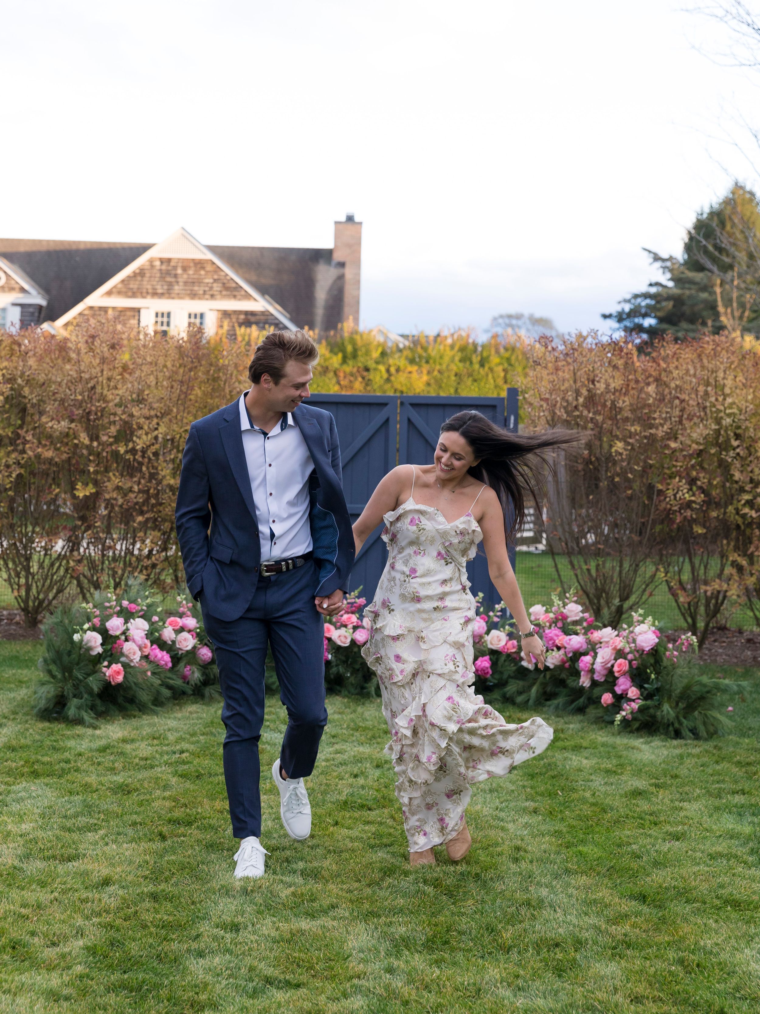 Joyful couple running through lush garden with pink roses, man in navy suit, woman in floral strapless gown.