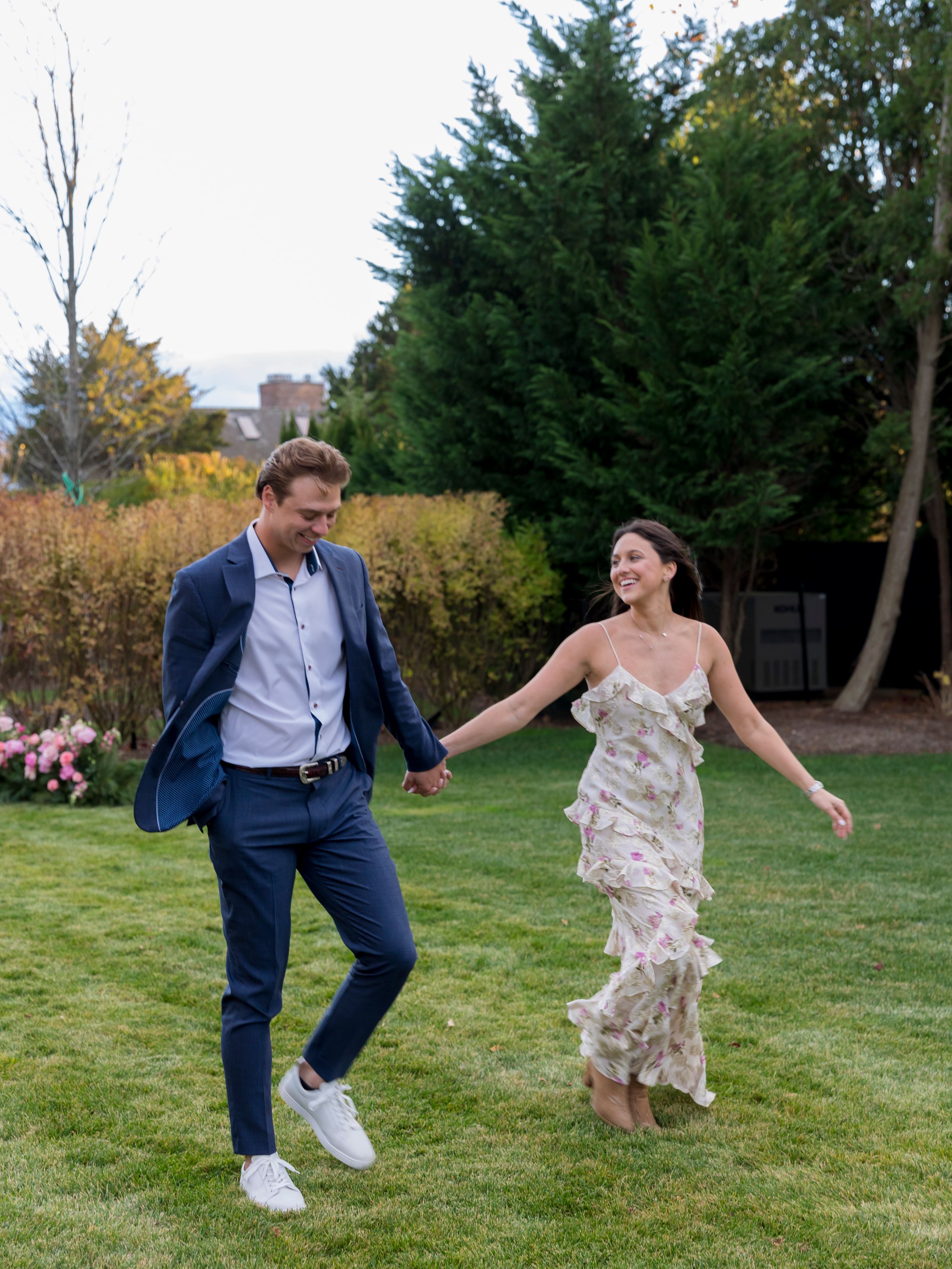 A joyful couple holds hands and runs across a lush green lawn, dressed in elegant semi-formal attire.