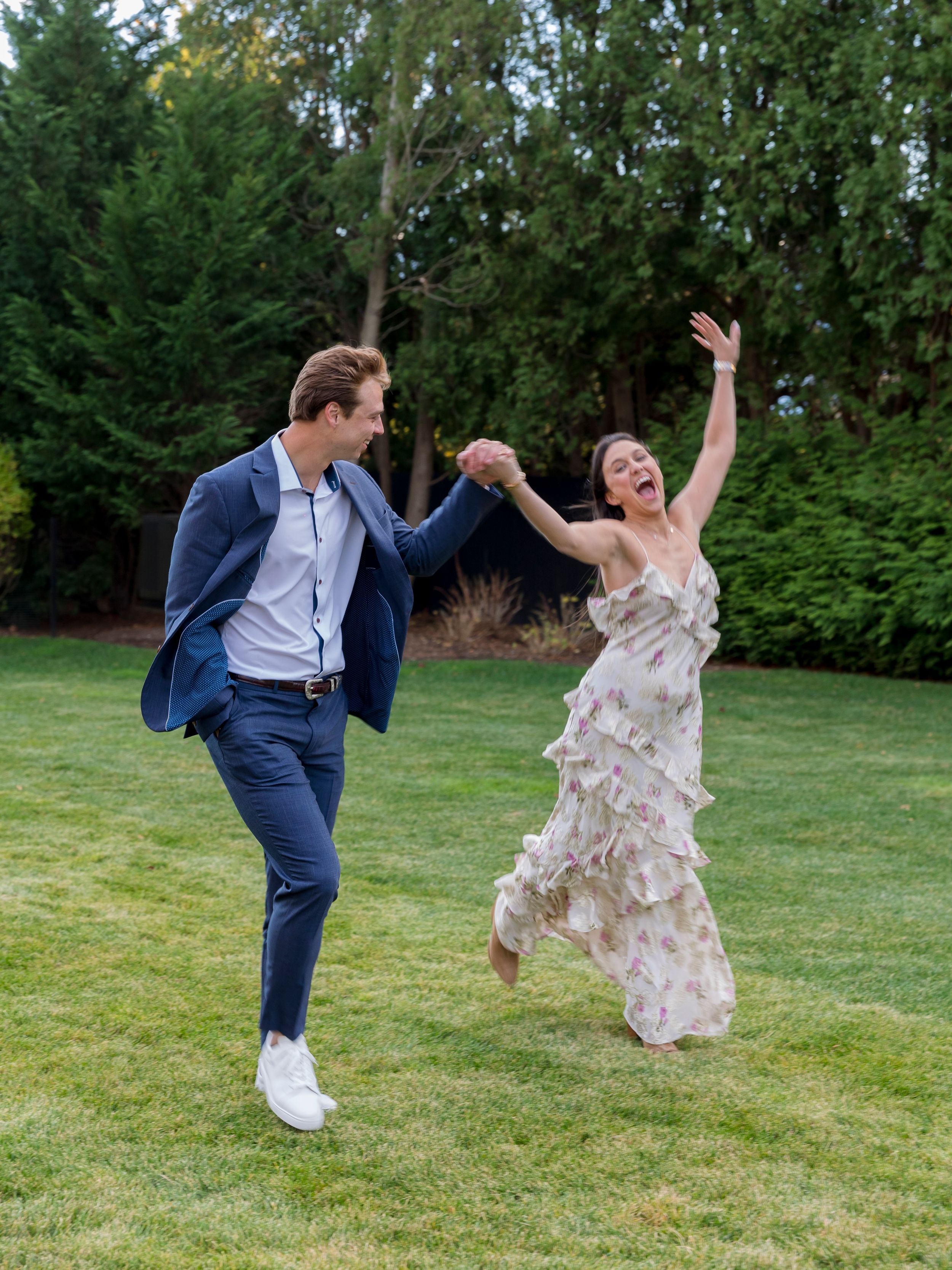 Happy couple dancing on green lawn, man in navy blazer holding hands with woman in ruffled floral dress.