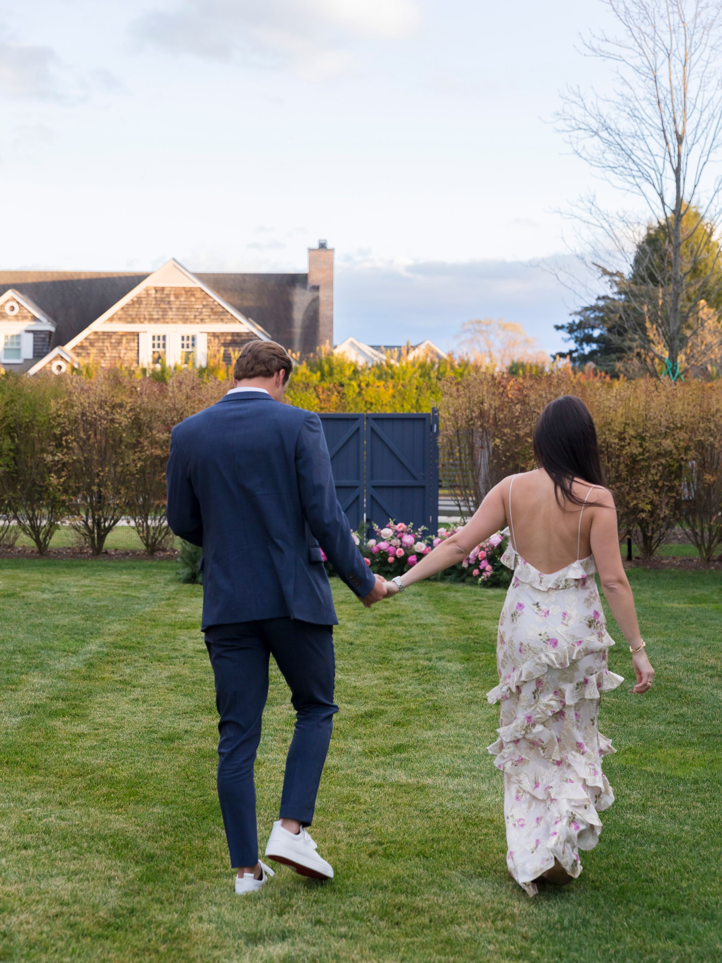 Couple holding hands walking across green lawn, man in navy suit, woman in backless floral ruffle gown, garden setting.