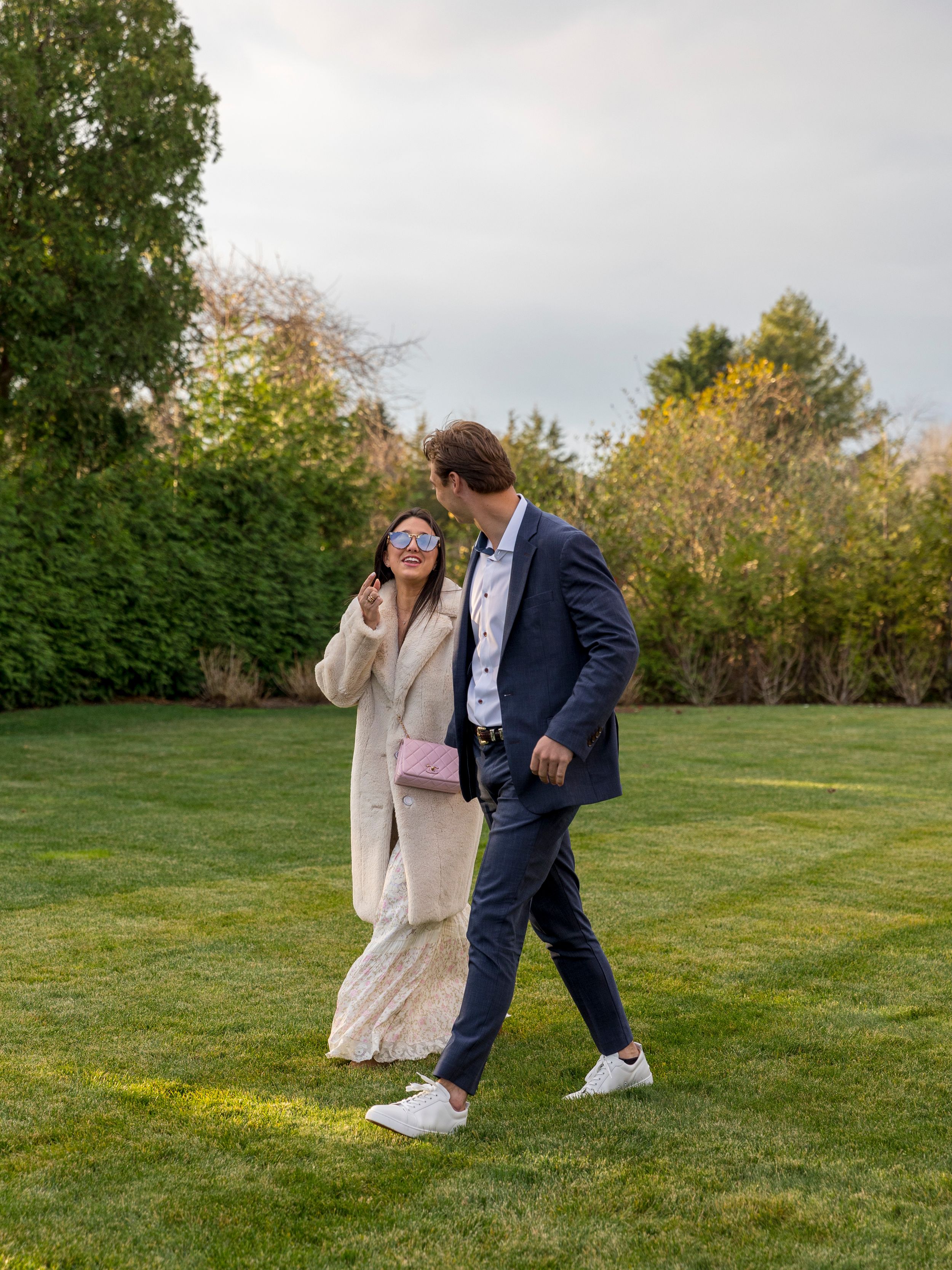 Couple walking on green lawn, woman in cream coat with pink bag, man in navy suit and white sneakers, lush garden backdrop.