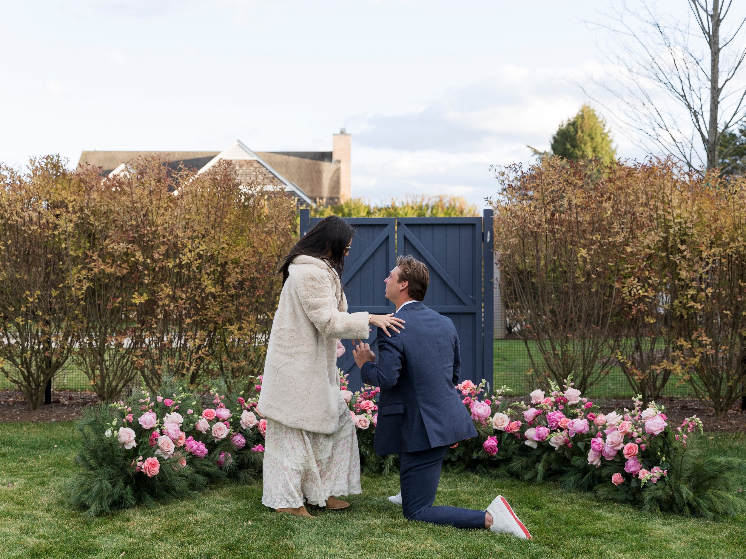 Man proposes on one knee to woman outdoors surrounded by pink floral arrangements near a blue garden gate.
