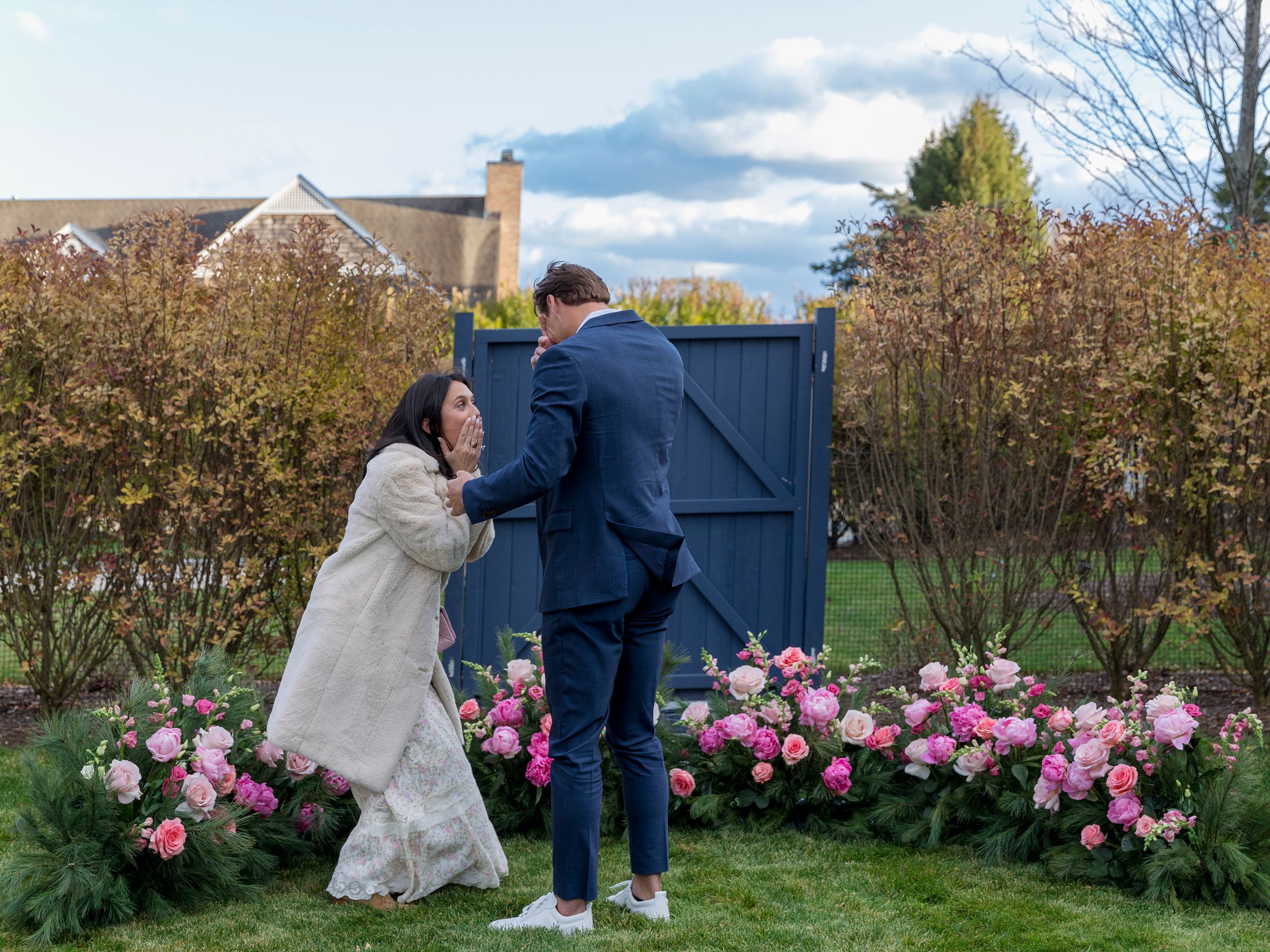 Couple sharing a romantic moment among pink floral arrangements in a garden with blue gate backdrop.