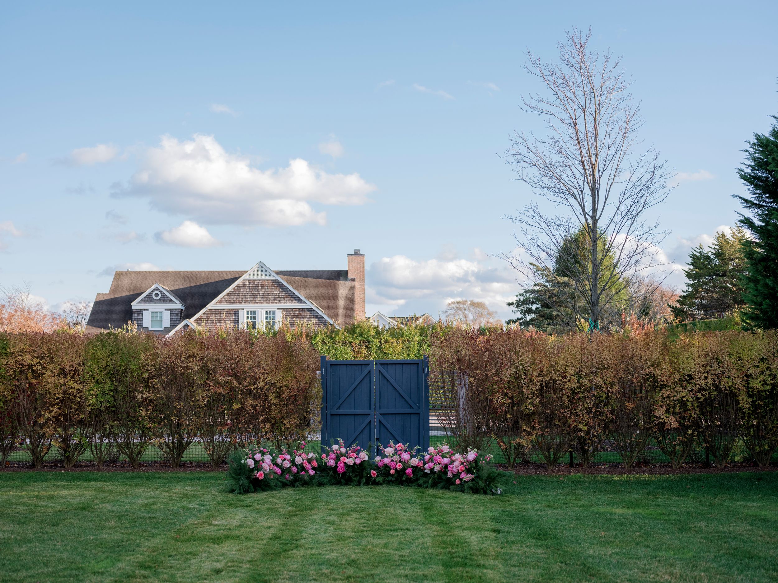 A lush green lawn with a blue gate surrounded by pink flowers and a hedge, with a charming house in the background.