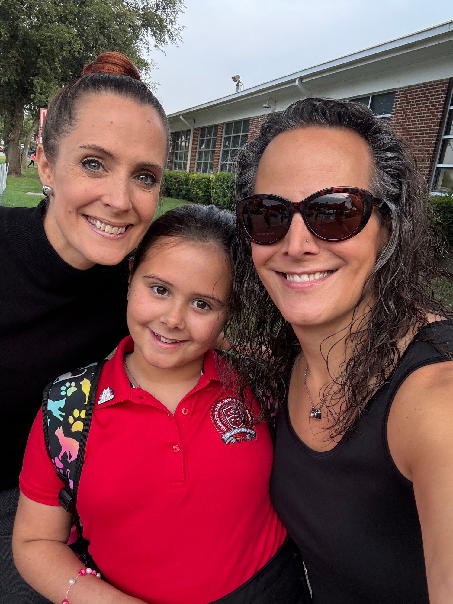 Two women and a young girl in a red polo smile together in a selfie outside a brick school building.  The first day of third grade.