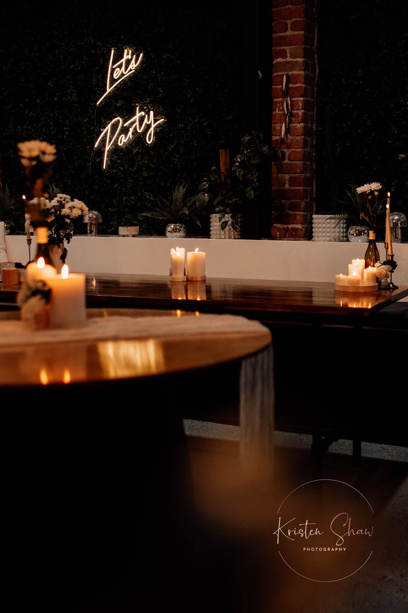 Dimly lit event space with neon 'Lets Party' sign, candles, and floral arrangements on dark tables.