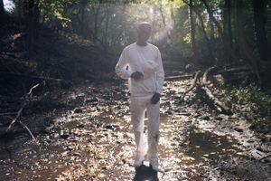 Man in white outfit walking through a sunlit forest path.