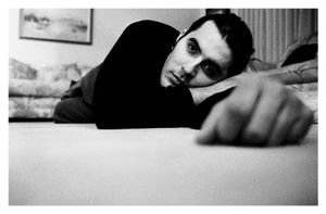 A young man lies on the floor resting his chin near a bed in black and white.