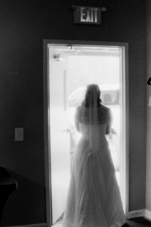 Silhouette of a bride in a wedding gown and veil standing in a bright doorway below an exit sign.
