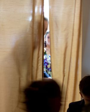 A young girl in a floral dress peeks through a gap in golden curtains at a wedding ceremony.