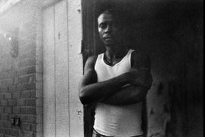 Young Black man in a white tank top leaning against a brick wall in a black and white photo.