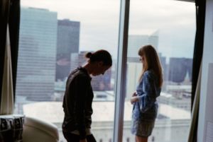 Two people standing in silhouette against large floor-to-ceiling windows with a city skyline backdrop.