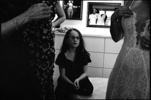 A young girl in glasses sits on a counter surrounded by women in formal attire.