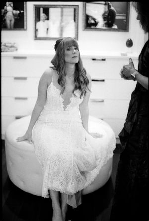 Bride in a lace white dress sits on a round ottoman during wedding preparations in black and white.