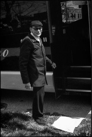 Black and white photo of an elderly man in a cap and coat standing by a bus.